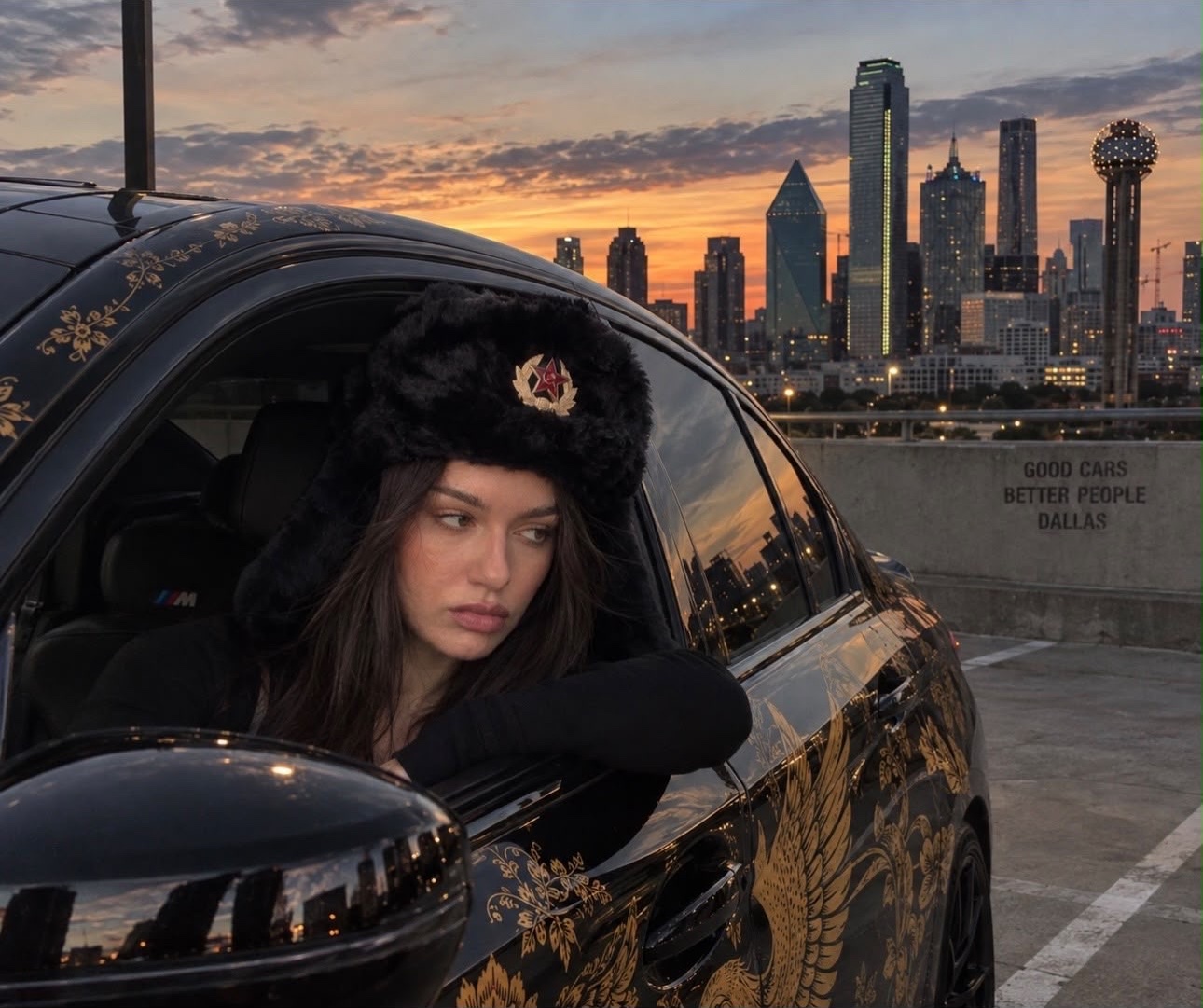 Katie in a fur hat looking out of a black BMW with gold Russian floral art on the door, Dallas skyline behind her