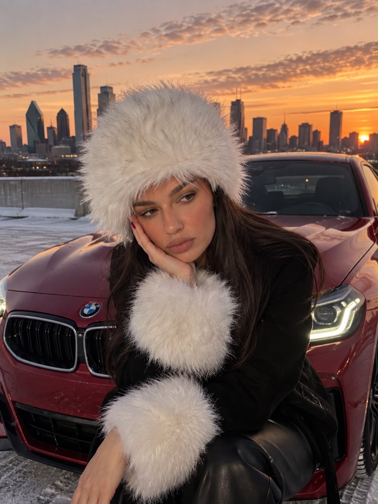 Katie in a white fur hat resting her cheek on her hand, with a red BMW and the Dallas skyline at sunset behind her